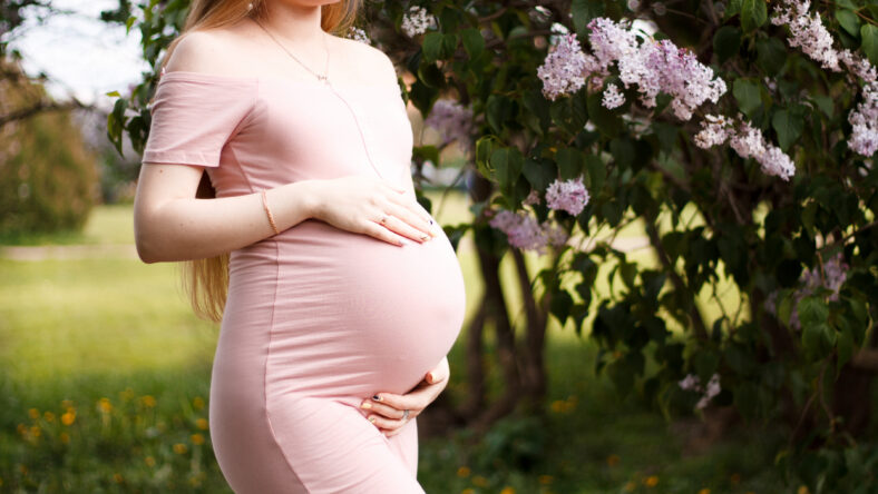 pregnant blonde girl in pink dress with bare shoulders holding hands belly with long hair on the background of lilacs and trees with a big belly on the eighth month of pregnancy close-up