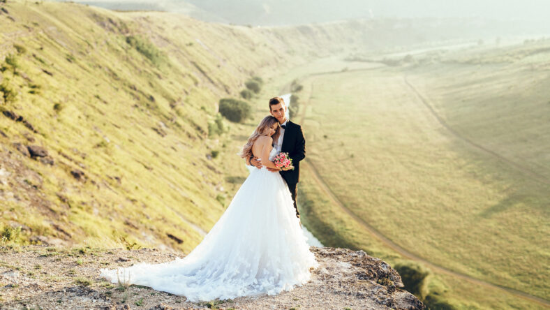 Full length body portrait of young bride and groom enjoying romantic moments outside at sunset in beautiful summer day. Wedding couple. Standing face to face with the green hills on background.