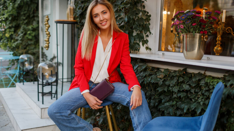 Young european confident woman with candid smile posing outdoor in the bar. Wearing red fashionable jacket.Traveling female enjoing vacations in the city.