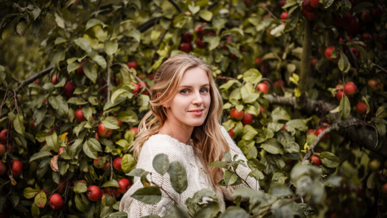 Natural outdoor portrait of blonde smiling woman in cozy sweater posing between branches full of red apples on a warm autumn evening