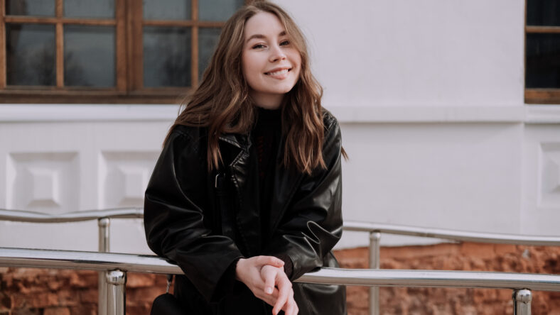Smiling young woman in black leather jacket standing in front of white wall, leaning on iron railings, looking at camera. Urban style and street fashion. Girl with curly hairstyle in casual outfit