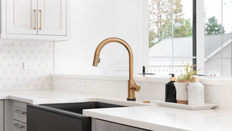 A beautiful sink in a remodeled modern farmhouse kitchen with a gold faucet, black apron or farmhouse sink, white granite, and a tiled backsplash.