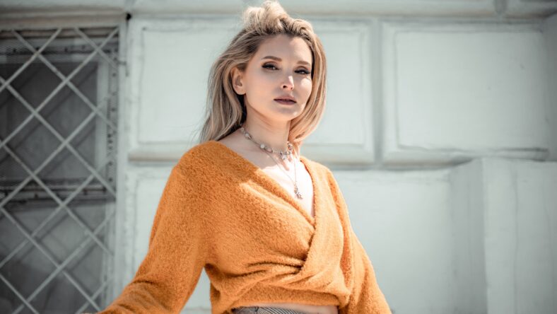A middle-aged woman looks like a good blonde with curly beautiful hair and makeup on the background of the building. She is wearing a yellow sweater