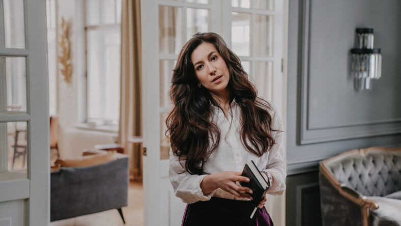 Calm brunette woman with wavy dark hair in white shirt and violet pants standing in living room, looking at camera, holding diary. Tired businesswoman at home after busy day at work. Business people.
