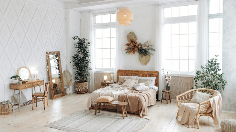 Cozy rustic bedroom with boho ethnic decor. Bright spacious apartment with large windows. Nobody. Wooden furniture. Boudoir table. Large mirror. Handmade textile. Plants in the interior.