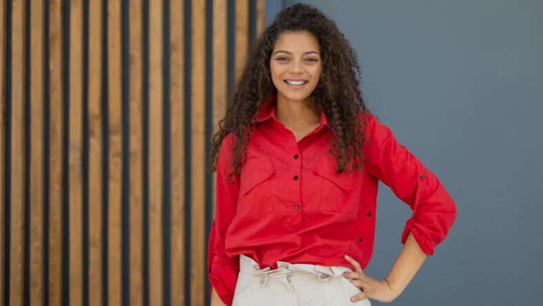 Young woman in red shirt standing against grey and wooden wall.