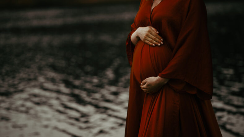 Woman in a beautiful red maternity evening dress by the water. Hope for a baby to come soon. Closeup shot of an unrecognizable pregnant woman.