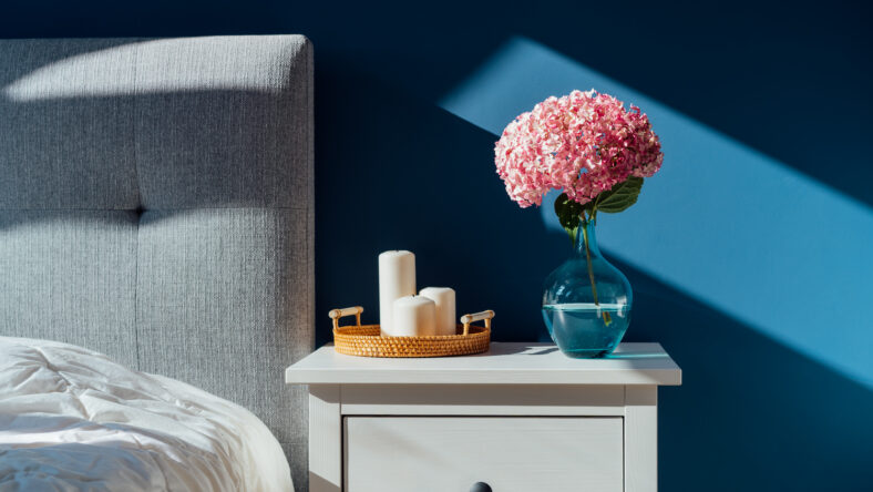 Stylish modern bedroom in dark colors. Cozy interior with navy blue walls, home decor. Bed with grey fabric headboard, white blanket, bedside table, vase with pink hydrangea flower, candles on tray