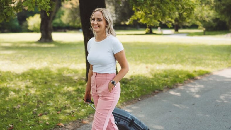 A young woman walking in a park with a suitcase. She has long blonde hair, wearing a white t-shirt and pink pants, smiling as she strolls along a path surrounded by greenery.